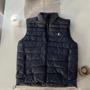 Mörkblå Polo Ralph Lauren väst - En mörkblå Ralph Lauren väst i storleken S som är i mycket bra skick. Det finns inga defekter. Den passar 175-185 cm. Den har burits ungefär 2-3 gånger, och det finns även qr-kod som man kan skanna för att säkerställa äkthet. Den är aningen blåare än vad som framkommer i bilderna. Hör av er vid övriga frågor!