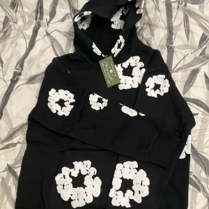 Svart hoodie med vita blommönster från Denim Tears - Säljer en svart hoodie från Denim Tears med stora vita blommönster tryckta över hela tröjan. Hoodien har en klassisk passform, huva och magficka. Tillverkad i USA och perfekt för dig som gillar streetwear.