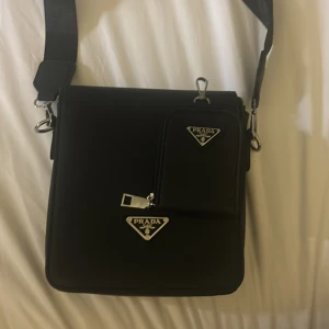 Svart axelväska från Prada - Säljer en svart axelväska från Prada med justerbar rem och silverfärgade detaljer. Väskan har en avtagbar liten ficka framtill med dragkedja och klassiska Prada-loggor i metall. Perfekt för att bära det viktigaste med stil.