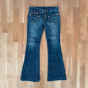 Blå bootcut jeans med fickdetaljer - Säljer ett par blå bootcut jeans med snygga slitningar och markerade sömmar. Jeansen har fram- och bakfickor med lock och knappdetaljer. Tillverkade i bomull med lite stretch för skön passform. Storlek M men skulle säga att den passar S bättre 