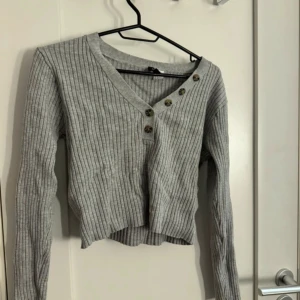 Grå ribbad croppad tröja med knappar - Säljer en grå ribbad croppad tröja med v-ringning och dekorativa knappar både vid halsen och på ärmarna. Tröjan har långa ärmar och är i ett mjukt, stretchigt material. Perfekt för lager-på-lager-stil.