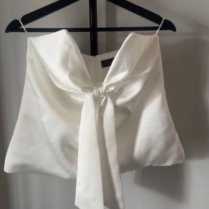 Vit axelbandslös topp med rosett - Säljer en elegant vit axelbandslös topp med stor rosettdetalj framtill. Toppen har en snygg omlottkänsla och är perfekt för dig som vill sticka ut. Tillverkad i ett slätt material som ger en lyxig känsla. Lappen är kvar!