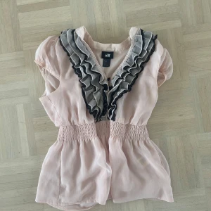 Ljusrosa blus med volang från H&M - Supersöt ljusrosa blus från H&M med korta puffärmar och volangdetaljer i grått och svart längs v-ringningen. Smock i midjan ger en fin passform. Perfekt för dig som gillar romantisk stil!