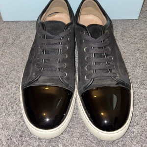 Svarta sneakers med lackad tå från Lanvin - Säljer ett par svarta sneakers från Lanvin med snygg lackad tå i svart och vita sulor. Skorna har snörning och ovandel i mocka, vilket ger en lyxig känsla. Perfekta för dig som gillar stilrena och unika detaljer.