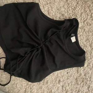 Svart ribbad croppad linnetopp med snörning - Säljer en svart ribbad croppad linnetopp från H&M med snygg snörningsdetalj framtill. Toppen är ärmlös och har en tight passform. 
