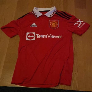 Manchester United hemmatröja 2022/23 - Röd Manchester United fotbollströja från Adidas med vit krage och svarta detaljer. Klubbmärke och Adidas-logga på bröstet, TeamViewer sponsortryck på framsidan och DXC på ärmen. Klassisk design med korta ärmar.
