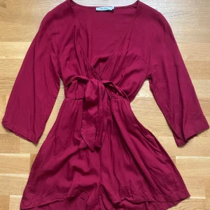 Vinröd playsuit/jumpsuit från NA-KD - Säljer en vinröd playsuit/jumpsut från NA-KD med trekvartsärm och knyt i midjan. Den har v-ringning och är i ett mjukt, lätt material. Storlek XS men skulle säga att den även passar för de som brukar ha storlek S.