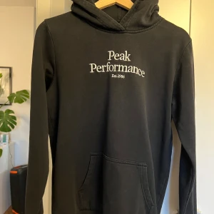 Svart hoodie från Peak Performance - Säljer en stilren svart hoodie från Peak Performance med broderad logga på bröstet. Tröjan har en klassisk känguruficka och en bekväm huva. Perfekt för en avslappnad stil.