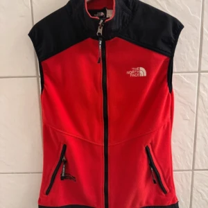 Röd fleeceväst från The North Face - Säljer en röd fleeceväst från The North Face med svarta detaljer och dragkedja. Västen har två praktiska fickor med dragkedjor och är tillverkad i polyester med Gore Windstopper-teknologi. Perfekt för utomhusaktiviteter!