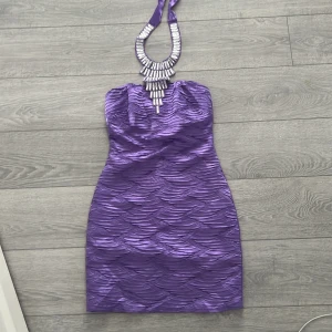 Lila axelbandslös klänning med smyckesdetalj - Snygg lila axelbandslös klänning med en unik smyckesdetalj runt halsen. Klänningen har en vågig textur som ger en elegant look. Perfekt för en kväll ute eller en speciell tillställning. Stl 36 turkisk motsvarar ungefär en 34