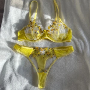 Gul blommig spets-bh och trosor - Säljer ett set med gul bh och matchande trosor i transparent material med blommiga broderier. Setet har justerbara axelband och en delikat spetsdetalj som ger en romantisk känsla. Perfekt för att känna sig extra fin. SJÄLVKLART OANVÄNT. 
