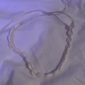 Vitt snäckhalsband - Säljer ett stilrent vitt halsband med snäckor. Perfekt för en bohemisk look eller sommarens stranddagar. Halsbandet har en justerbar snörning för att passa olika storlekar. Enkelt men elegant tillägg till din smyckessamling.