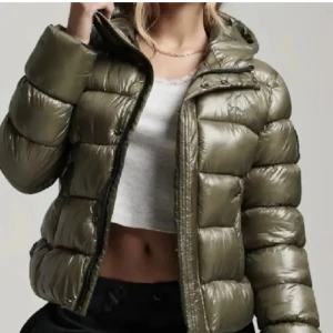 pufferjacka - Snygg olivgrön pufferjacka med dragkedja. Jackan har en glansig yta och är perfekt för kyligare dagar. Den har långa ärmar och en hög krage för extra värme.