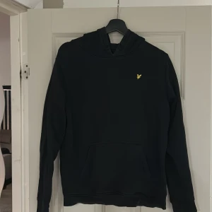 Svart hoodie från Lyle & Scott - Säljer en stilren svart hoodie från Lyle & Scott med en liten gul logga på bröstet. Tröjan har en klassisk design med huva och långa ärmar. Perfekt för en avslappnad stil.