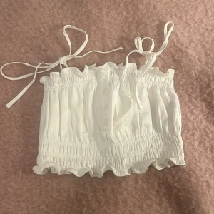 Vit axelbandslös topp med knytband - Säljer en söt vit axelbandslös topp med knytband och knappar framtill. Toppen har en design med volangdetaljer vid kanterna. Perfekt för varma sommardagar! Man kan helt skjustera storleken på banden och toppen är super stretchig och bekväm 💗💗ny pris 1200kr