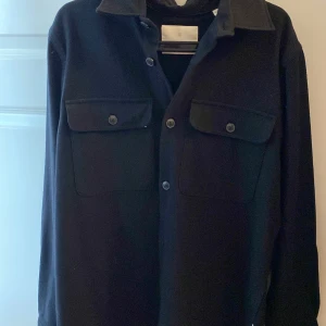 Svart overshirt från Jack & Jones - Snygg svart overshirt från Jack & Jones med knappar framtill och två bröstfickor med lock. Perfekt för lager-på-lager-stil och passar till många olika outfits. Den är ganska tjock och varm, men också ganska relativt stor i storleken.