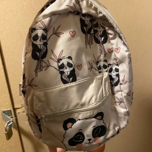 Beige ryggsäck med pandamönster - Säljer en söt beige ryggsäck med pandamönster och små hjärtan. Väskan har en praktisk dragkedja framtill och justerbara axelremmar i rosa. Perfekt för att bära dina dagliga nödvändigheter med stil 🐼💕