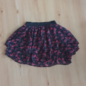 Blommig kortkjol med volang - Söt blommig kortkjol med volangdetaljer. Kjolen har en elastisk midja och ett härligt mönster i rött på en svart bakgrund. Perfekt för en somrig look!