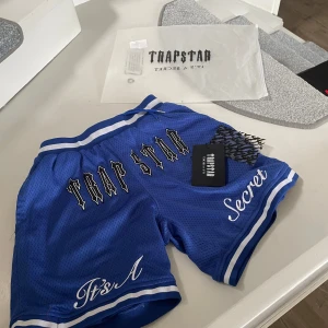 Blåa trapstar shorts - Snygga blå shorts från Trapstar med texten 'Trapstar' och 'It's A Secret' tryckt på framsidan. De har en elastisk midja med snörning och vita detaljer längs kanterna. Perfekta för en avslappnad stil.
