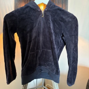Juicy couture kofta  - Säljer en snygg mörkblå hoodie med dragkedja och huva. Den har ett stort, dekorativt JC-märke i guld på ryggen. Perfekt för en avslappnad stil.