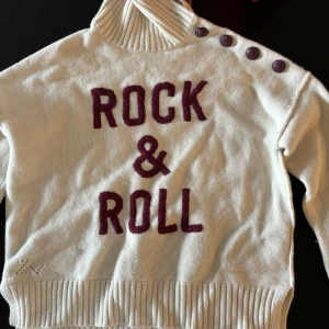 Zadig&voltaire tröja  - Säljer en vit stickad tröja med texten 'Rock & Roll' i vinrött på framsidan. Tröjan har en hög krage och dekorativa knappar på axeln. Perfekt för en cool och avslappnad stil.