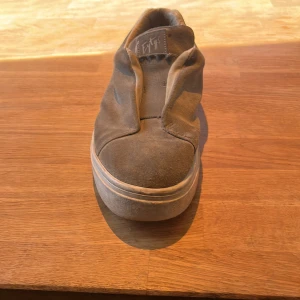 Gråa  slip-on sneakers - Säljer ett par gråa slip-on sneakers med rund tå och en diskret lila detalj på hälen. Skorna har en bekväm och stilren design utan snörning, perfekt för en avslappnad look. Synligt använda finns ej låda