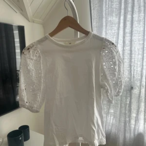 Vit blus med spetsärmar från H&M - Säljer en söt vit blus från H&M med korta spetsärmar. Blusen har ett fint blommigt spetsmönster på ärmarna som ger en romantisk touch. Perfekt för vår och sommar! 🌸