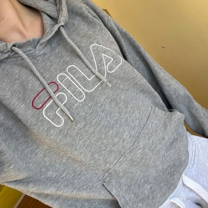 Grå hoodie från FILA - Säljer en grå hoodie från FILA med en stor logga i vitt och rött på bröstet. Tröjan har en bekväm passform med långa ärmar och en praktisk magficka. Perfekt för en avslappnad stil. Hör av dig vid intresse! 😊