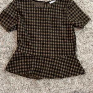 Rutig topp med volangdetalj - Säljer en snygg rutig topp i brunt och svart med korta ärmar och volangdetalj vid midjan. Toppen har en dragkedja i ryggen för enkel på- och avtagning. Perfekt för en stilren look! Från Zara