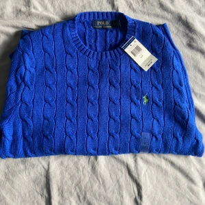 Blå stickad tröja från Ralph Lauren - Säljer en snygg blå stickad tröja från Ralph Lauren med kabelstickat mönster. Tröjan har en broderad grön logotyp på bröstet och rund halsringning. Perfekt för en stilren look! Har aldrig använts!