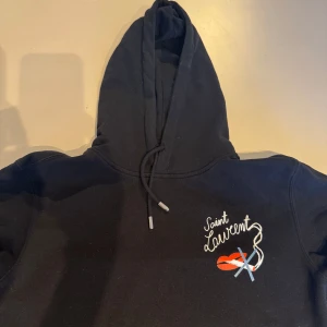 Svart hoodie från Saint Laurent - Snygg svart hoodie från Saint Laurent med broderad logga och läppmotiv på bröstet. Tröjan har en klassisk huva med dragsko och långa ärmar. Perfekt för en stilren och avslappnad look.