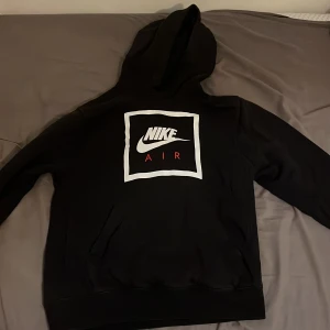 Svart hoodie från Nike - Säljer en svart hoodie från Nike med en stor vit och röd logga på framsidan. Tröjan har en klassisk känguruficka och en bekväm huva. Perfekt för en avslappnad stil.