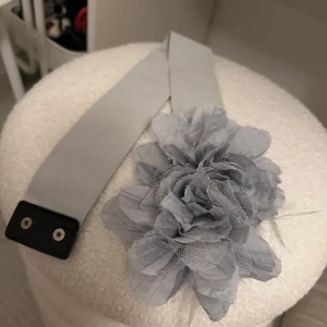Ljusblå bälte med blommig detalj - Snyggt grått bälte med en stor blommig detalj i tyg och nät. Perfekt för att ge en extra touch till din outfit. Bältet har en enkel knäppning med tryckknappar.