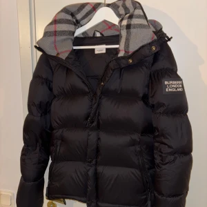 Svart dunjacka från Burberry - Snygg svart dunjacka från Burberry med en klassisk huva. Jackan har en dragkedja framtill och långa ärmar. Perfekt för kyliga dagar med stil. jackan kan även bli till en väst bara genom dragkedjan