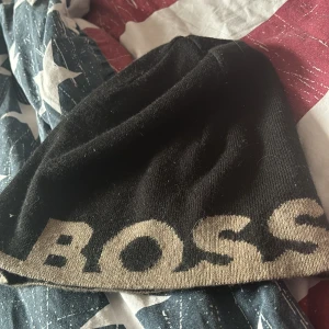 Svart mössa från Boss - Snygg svart mössa från Boss med stor logga i beige. Perfekt för att hålla värmen med stil. Tillverkad i mjukt material för en bekväm passform.