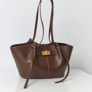 Brun axelväska med gulddetaljer - Snygg brun axelväska i skinn med eleganta gulddetaljer. Väskan har långa remmar och en stilren design med ett dekorativt spänne framtill. Perfekt för att bära dina dagliga nödvändigheter med stil.