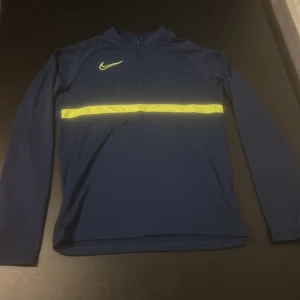 Blå långärmad tröja från Nike - Säljer en blå långärmad tröja från Nike med en rand över bröstet och en dragkedja vid halsen. Tröjan är tillverkad i Dri-FIT material, perfekt för träning.