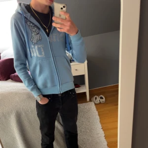 Ljusblå hoodie från Vonstudio - Säljer en ljusblå hoodie från Vonstudio med dragkedja och ett glittrigt tryck på framsidan. Tröjan har en mysig huva och praktiska fickor framtill. Perfekt för en avslappnad stil. Säljer då den är för liten. Skicket är 10/10 utan några defekter. Priset är inte hugget i sten - kontakta vid minsta fundering. 