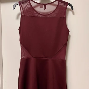 Vinröd klänning med meshdetaljer - Snygg vinröd klänning med meshdetaljer upptill. Den är ärmlös och har en elegant design som passar perfekt för festliga tillfällen. Klänningen har en figurnära passform och är gjord i ett mjukt material.