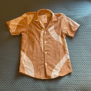 Beige kortärmad skjorta från OAS - Säljer en snygg beige kortärmad skjorta från OAS i mjuk frotté. Skjortan har ett unikt mönster i vitt och knäpps med knappar framtill. Perfekt för en avslappnad stil. Nypris 1300kr