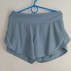 Blå träningsshorts från Lululemon - Säljer ett par blå träningsshorts från Lululemon. Shortsen har en diskret logga baktill och praktiska fickor med dragkedja. Perfekta för träning och sommaraktiviteter. 