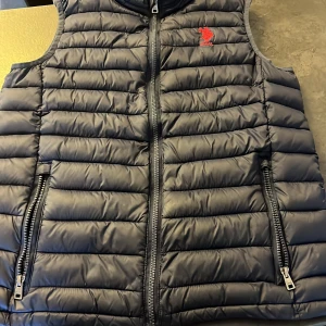 Mörkblå dunväst från U.S. Polo Assn. - Snygg mörkblå dunväst från U.S. Polo Assn. med dragkedja och broderad logga på bröstet. Perfekt för kyliga dagar med sin quiltade design och praktiska fickor med dragkedja.