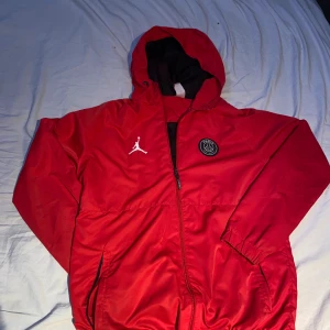 Röd vindjacka från Jordan x PSG - Säljer en snygg röd vindjacka från Jordan i samarbete med Paris Saint-Germain. Jackan har en dragkedja framtill, huva och PSG-logga på bröstet. Perfekt för sportiga tillfällen eller som en stilren vardagsjacka. Finns en liten fläck på ena armen därav priset.