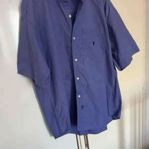 Ralph Lauren  - Säljer en klassisk blå skjorta från Polo i 100% bomull. Skjortan har korta ärmar och en bröstficka med en liten broderad logotyp. Perfekt för en stilren look.