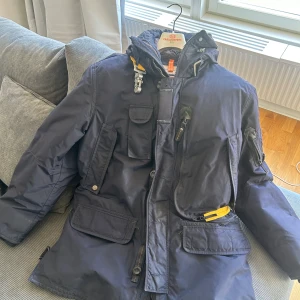 Mörkblå parkajacka från Parajumpers - Säljer en snygg mörkblå parkajacka från Parajumpers. Jackan har flera praktiska fickor, en avtagbar huva och en quiltad insida för extra värme. Den har också en gul detalj vid kragen och en pälsdetalj som kan fästas på huvan. (Kvitto finns) Orginal galge med följer. Inköpspris 10700kr 