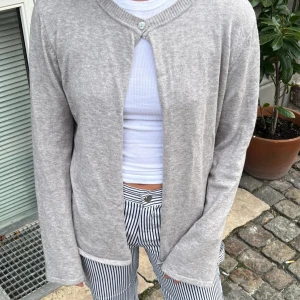 Grå kofta från design by si  - Säljer en stilren grå kofta med en enkel knappdetalj vid halsen. Koftan har långa ärmar och en mjuk, bekväm passform. Perfekt för lager-på-lager-stil. Passar till både jeans och kjol.