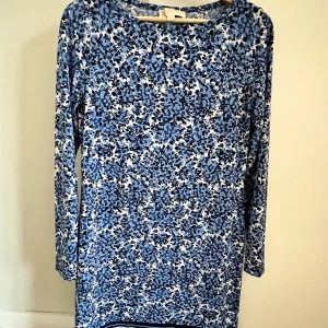 Blommig klänning från Michael Kors - Snygg blommig klänning i blå och vit från Michael Kors. Klänningen har långa ärmar och en rund halsringning. Perfekt för en stilren look med sitt vackra mönster och mjuka material. Slimfit modell så fin