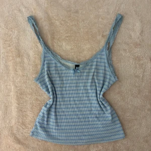 Blå randig linnetopp från H&M Divided - Säljer en söt blå och vit randig linnetopp från H&M Divided. Toppen har spetsdetaljer vid axelbanden och en liten rosett framtill. Perfekt för varma sommardagar! 💙