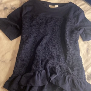 Mörkblå topp med volangdetalj - Säljer en mörkblå topp med korta ärmar och en fin volangdetalj vid nederkanten. Toppen har en smockad textur på framsidan som ger en unik look. Perfekt för att piffa upp vardagsstilen!