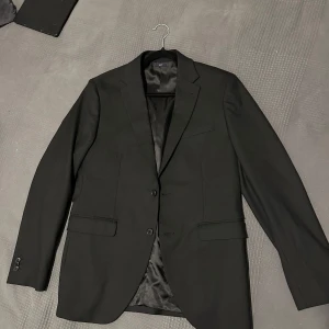 Svart kostym med vit skjorta från Riley - Elegant svart kostym från Riley med matchande kavaj och byxor. Kavajen har klassisk design med två knappar och fickor. Den vita skjortan är stilren och passar perfekt till kostymen. Kostymen är Slimfit, kavajen och skjortan är M storlek. Byxorna är 100 cm i längd och 45 cm i bredd. Kostymen är använd en gång. Säljs på grund av att den inte passar längre då jag har ökat i vikt. Passform: EU 48 passar 180-185cm